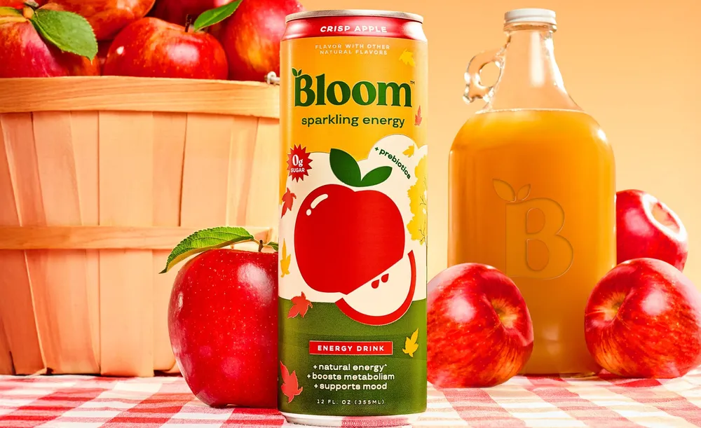 Bloom Sparkling Energy Crisp Apple can beside a glass jug of amber juice, red apples and an apple basket