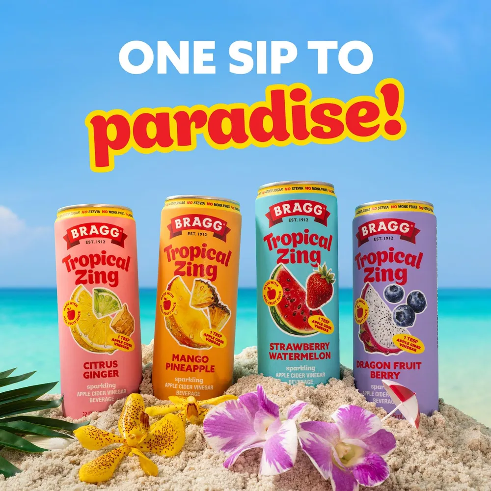 Four Bragg Tropical Zing sparkling apple cider vinegar cans are arranged in sand against a bright beach-and-sky backdrop, with tropical flowers and leaves in the foreground
