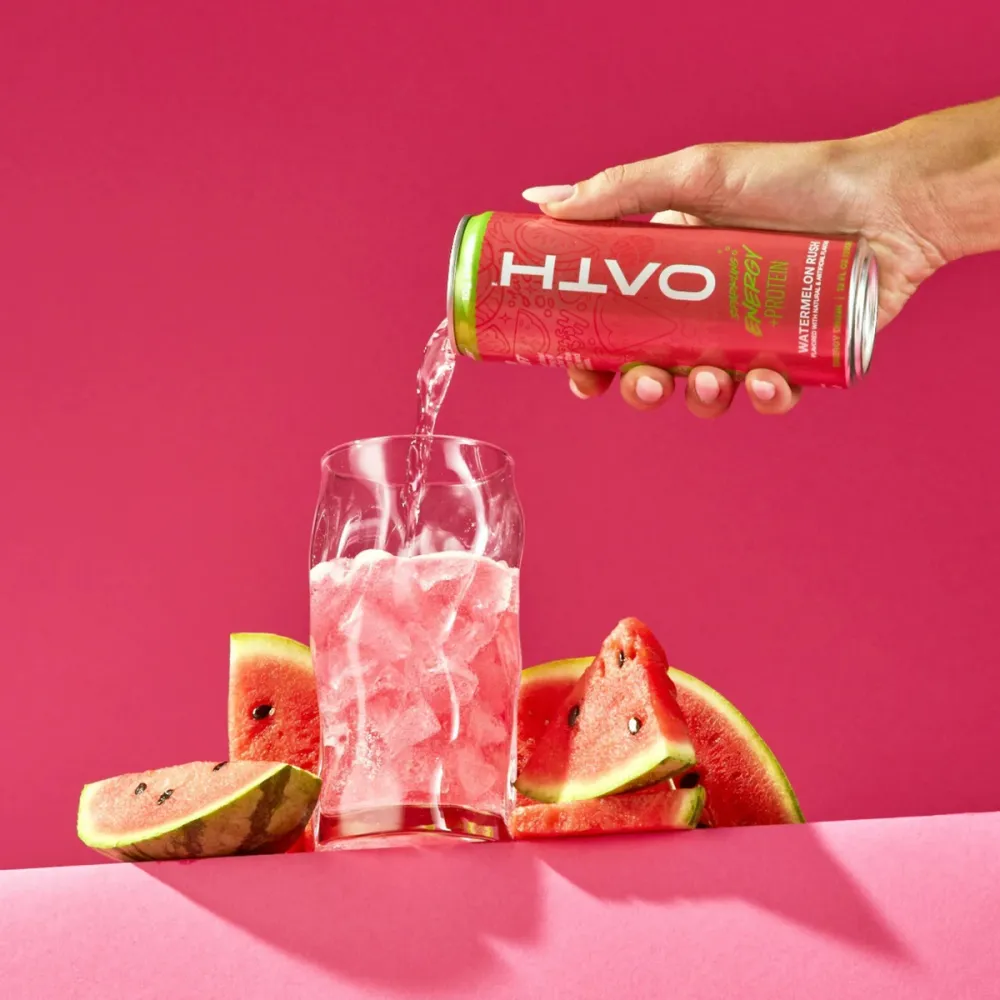 A hand pours a red OATH can into an ice-filled glass against a bright pink background