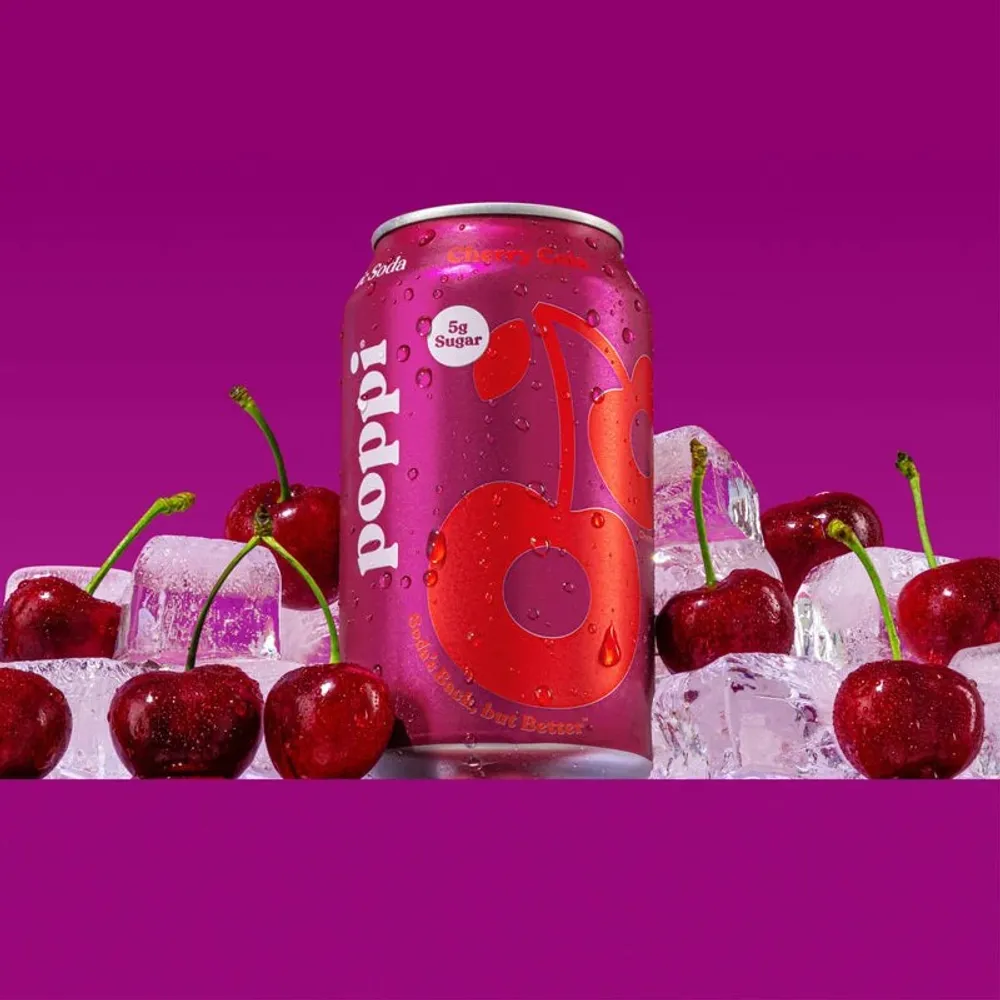 A pink Poppi can labeled “Cherry Cola” stands among red cherries and ice against a bright magenta background