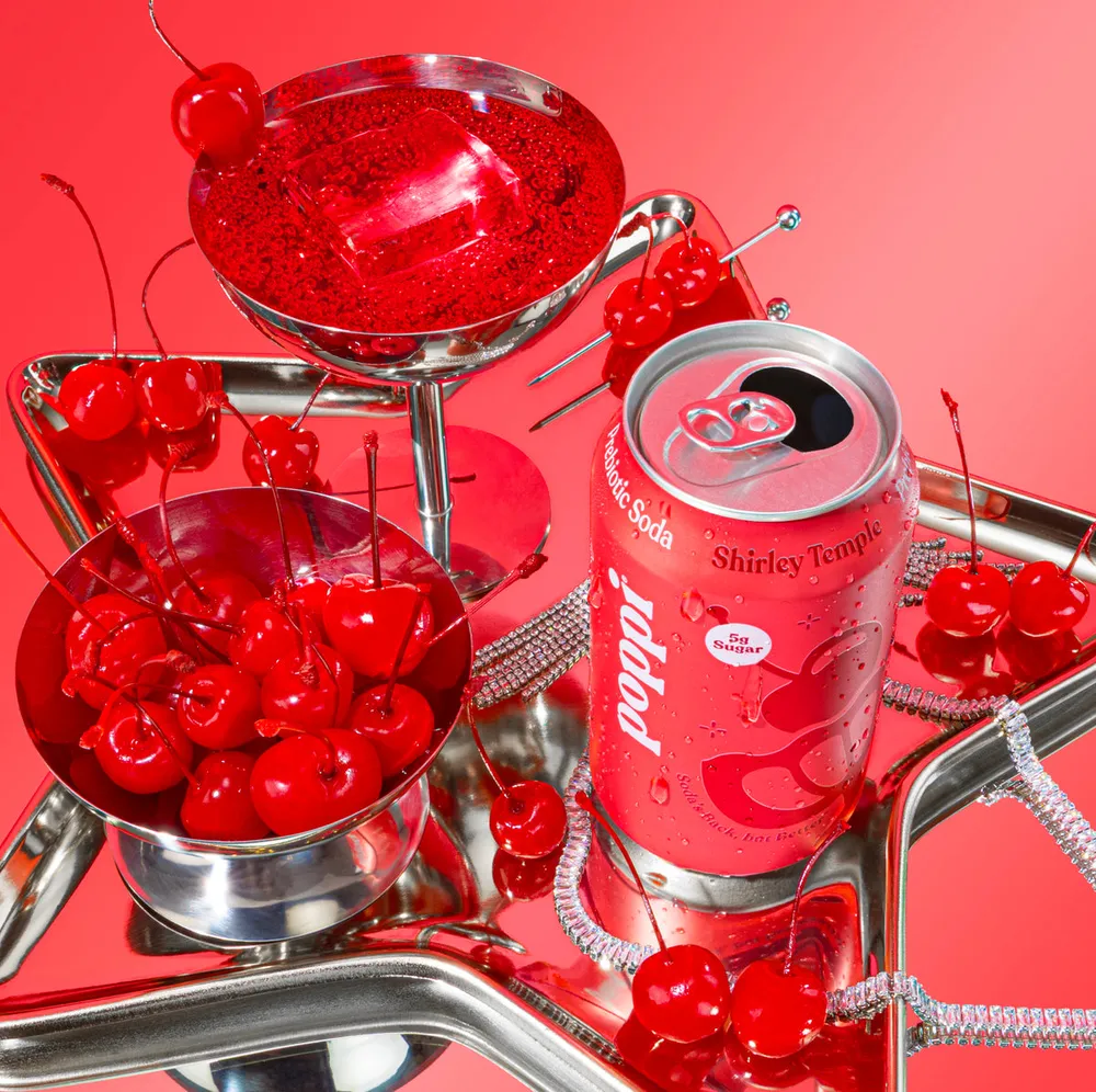 A pink Poppi Shirley Temple soda can sits among bowls of bright red cherries and a red cocktail in a shiny metal display