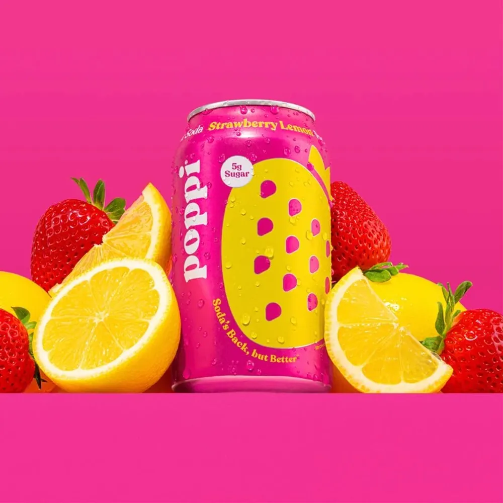 A pink and yellow Poppi Strawberry Lemon can stands between fresh strawberries and halved lemons against a hot pink background
