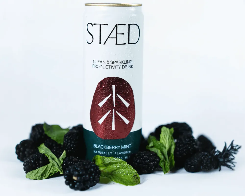 A white Staed Clean & Sparkling Productivity Drink can stands upright against a light background, surrounded by blackberries and mint leaves