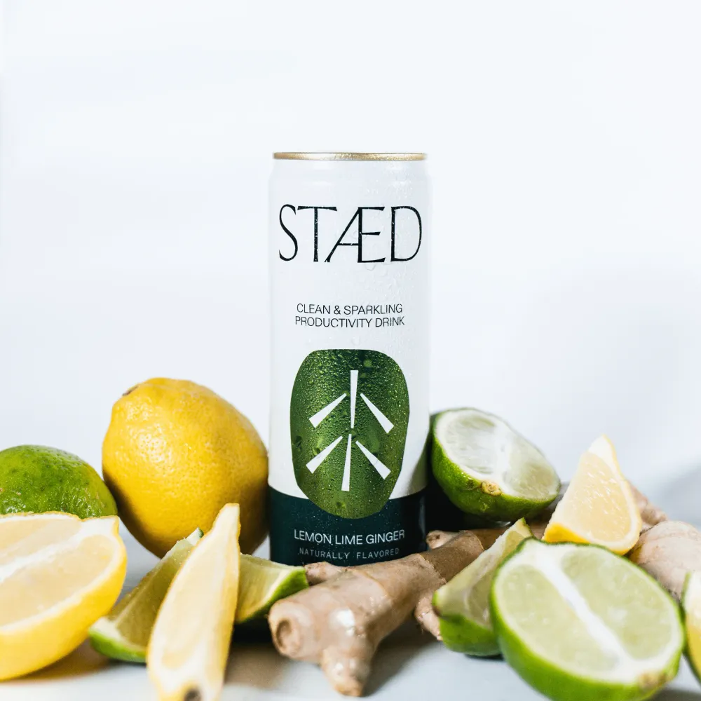A white Staed Clean & Sparkling Productivity Drink can stands upright among lemons, limes, citrus wedges, and a piece of ginger on a pale surface