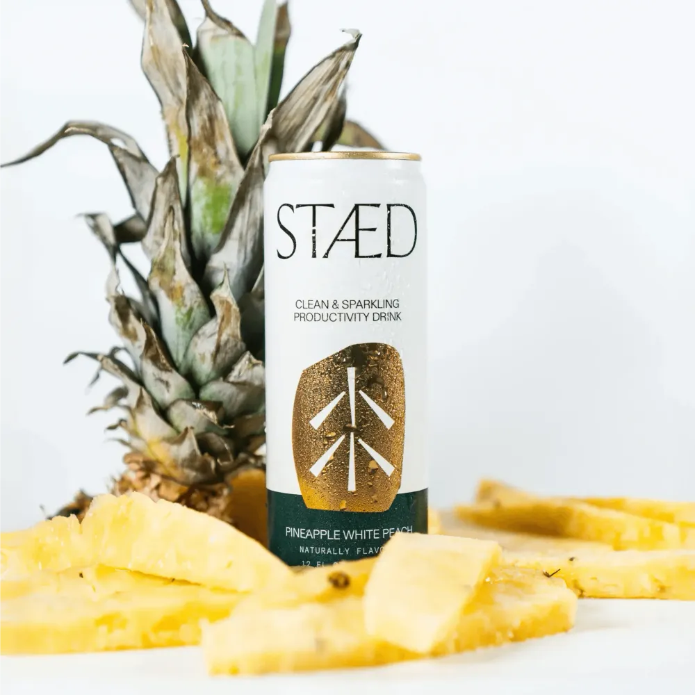 A white Staed Clean & Sparkling Productivity Drink can labeled Pineapple White Peach stands among pineapple leaves and fresh pineapple slices